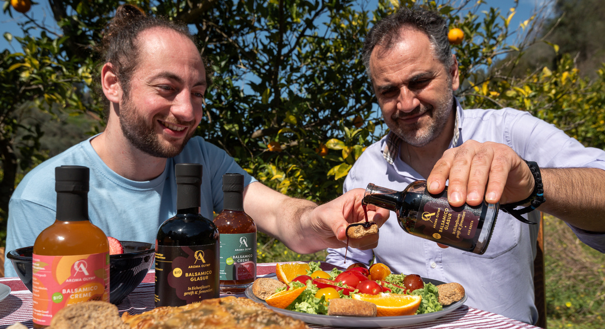 KI generiert: Zwei Männer genießen ein Essen im Freien mit Salat und Balsamico. Im Hintergrund sind Orangenbäume zu sehen.