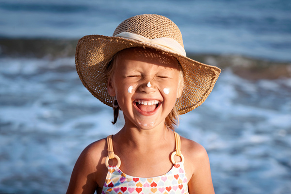 KI generiert: Ein lachendes Kind am Strand mit Sonnenhut und Sonnencreme auf dem Gesicht.