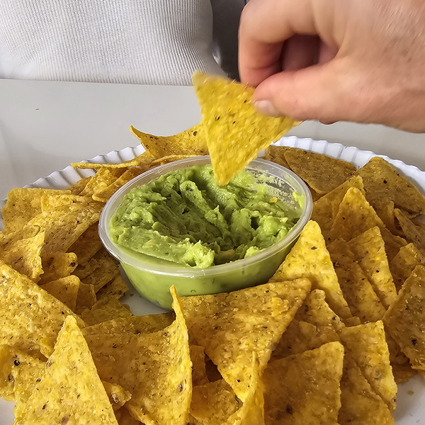 KI generiert: Das Bild zeigt eine Hand, die einen Tortilla-Chip in eine Schale mit Guacamole taucht. Rundherum liegen zahlreiche weitere Tortilla-Chips auf einem Teller.