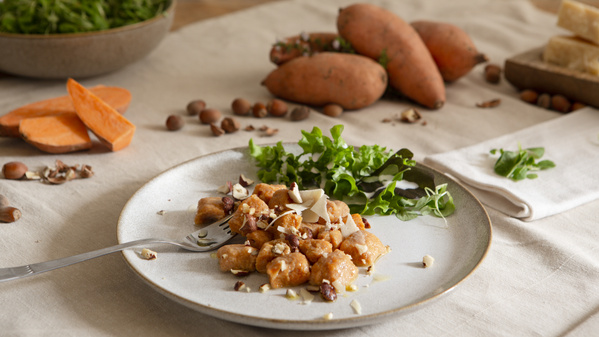 KI generiert: Ein Teller mit Gnocchi, Nüssen und Rucola, Süßkartoffeln im Hintergrund.