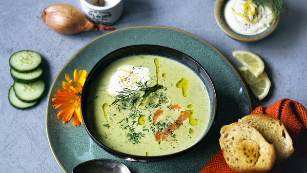 Ein Teller mit grüner Suppe, garniert mit Dill und Creme, umgeben von Brot, Zitrone und Gurken.