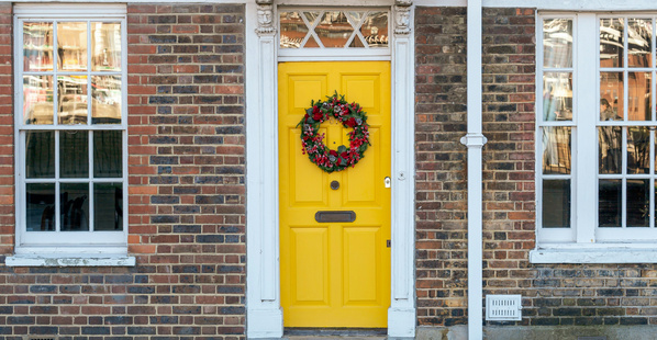 Das Bild zeigt eine gelbe Tür eines Backsteinhauses, die mit einem dekorativen Kranz geschmückt ist. Die Umgebung ist ruhig und die Fassade wirkt einladend.