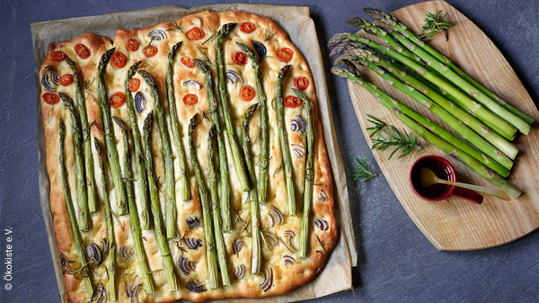 KI generiert: Eine Spargel-Focaccia auf Backpapier neben frischem Spargel und Olivenöl. Text: "ökokiste e.V."