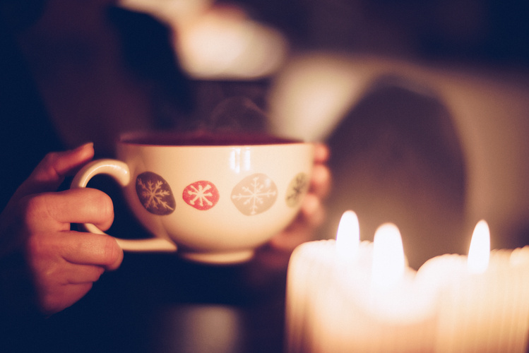 KI generiert: Eine Hand hält eine winterlich dekorierte Tasse, Kerzen im Vordergrund.