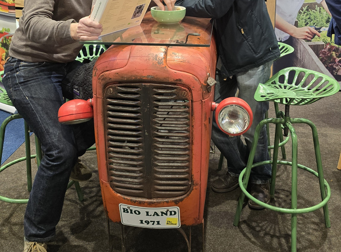 Alter kleiner roter Trecker mit einem herabhängenden  Scheinwerfer als Tisch umfunktioniert, daneben ein Treckersitz-Stehstuhl