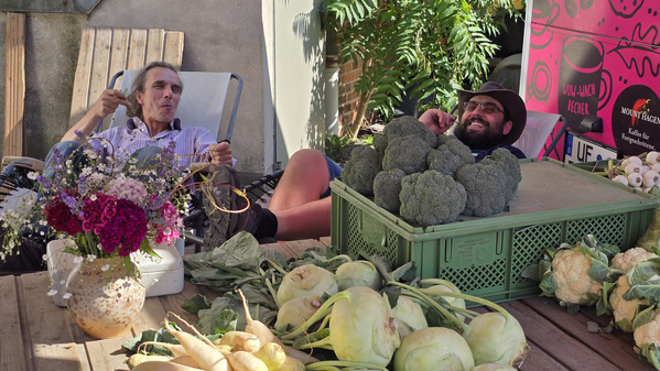 KI generiert: Zwei Männer entspannen sich hinter einem Tisch mit Gemüse und Blumen.