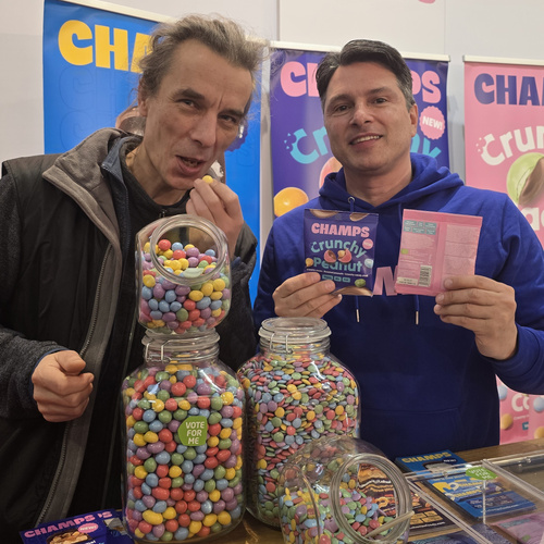 KI generiert: Zwei Männer stehen vor Gläsern mit bunten Süßigkeiten und halten eine Packung "CHAMPS Crunchy".