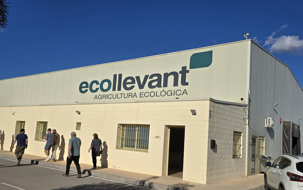 KI generiert: Das Bild zeigt ein Gebäude mit der Aufschrift "ecollevant Agricultura Ecológica" auf der Fassade. Vor dem Gebäude gehen mehrere Personen auf einem Bürgersteig.