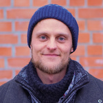 KI generiert: Ein Mann in Winterkleidung, bestehend aus Mütze und Schal, steht vor einer Ziegelwand und blickt in die Kamera. Der Hintergrund ist eine unscharfe Ziegelmauer.