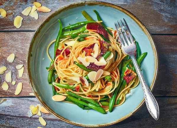 KI generiert: Ein Teller Spaghetti mit grünen Bohnen, getrockneten Tomaten und Mandelblättchen, auf einem Holztisch.