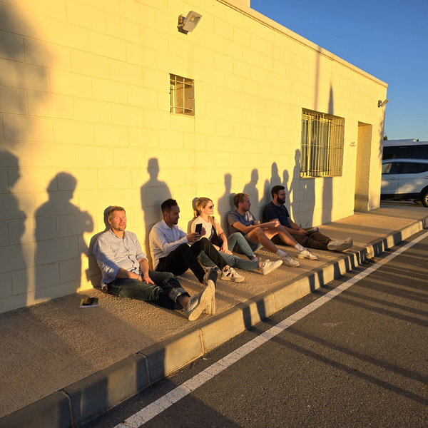 KI generiert: Eine Gruppe von Menschen sitzt in der Sonne an einer Wand, während ihre langen Schatten auf die Wand fallen. Sie entspannen sich auf einem Gehsteig neben einer Straße.