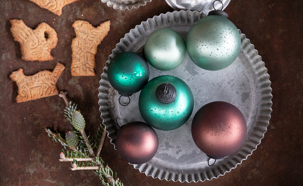 Das Bild zeigt eine Sammlung von Weihnachtskugeln in verschiedenen Grüntönen auf einem metallischen Tablett, daneben liegen Plätzchen in Tierform. Ergänzt wird die Szene durch einige Tannenzweige.