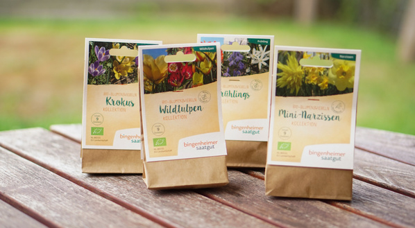 KI generiert: Das Bild zeigt vier Papiertüten mit Blumenzwiebeln von Bingenheimer Saatgut, die auf einem Holztisch stehen. Die Tüten sind mit verschiedenen Blumensorten wie Krokussen, Wildtulpen, Frühlingsblumen und Mini-Narzissen beschriftet.