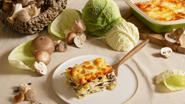 Eine Lasagne auf einem Teller, umgeben von frischen Pilzen und Wirsingblättern.