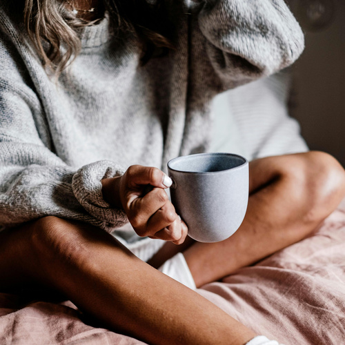 KI generiert: Eine Person hält eine graue Tasse und sitzt gemütlich in einem Pullover auf dem Bett.