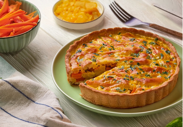 KI generiert: Gemüsequiche auf einem Teller, umgeben von Schalen mit rohen Karotten und geschnittenem Gemüse.