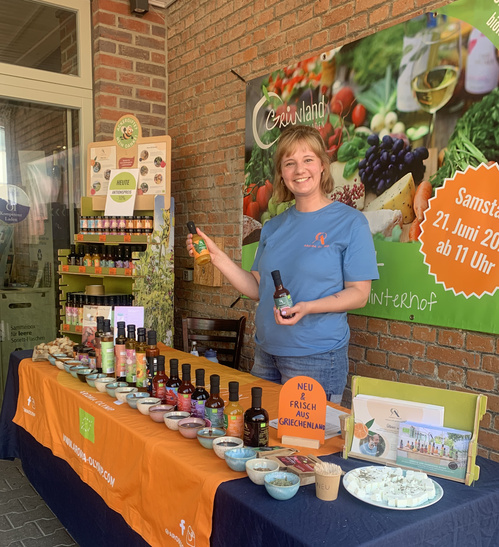KI generiert: Eine Frau steht an einem Verkaufsstand mit griechischen Produkten, im Hintergrund ein Banner von Grünland.