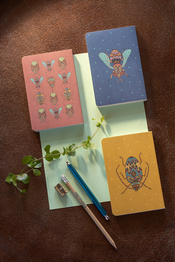 KI generiert: Drei farbige Notizbücher mit Insektenmuster, zwei Stifte, ein Spitzer und grüne Blätter auf einem Tisch.