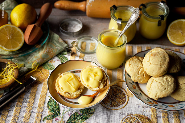 Auf 2 Tellern sind einmal aufgeschnittene Scones mit Lemoncurds und ein Stapel Lemoncurds. Dahinter stehen 1 geöffnetes und weitere Gläser von dem lemon Curd, umgeben von den Zutaten auf einer Decke mit Zitronenmotiv.