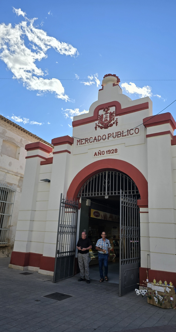 KI generiert: Das Bild zeigt den Eingang eines öffentlichen Marktes mit der Inschrift "Mercado Publico Año 1928" über dem Torbogen. Zwei Personen sind zu sehen, die den Markt verlassen.