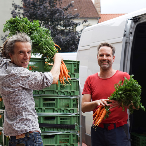 Zwei Männer lächeln, während sie Karotten vor einem Lieferwagen halten. Im Hintergrund sind grüne Gemüsekisten gestapelt.