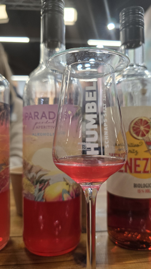 KI generiert: Ein Glas mit rotem Getränk vor Flaschen. Text auf dem Glas: "HUMBEL SCHNAPS SEIT 1918".