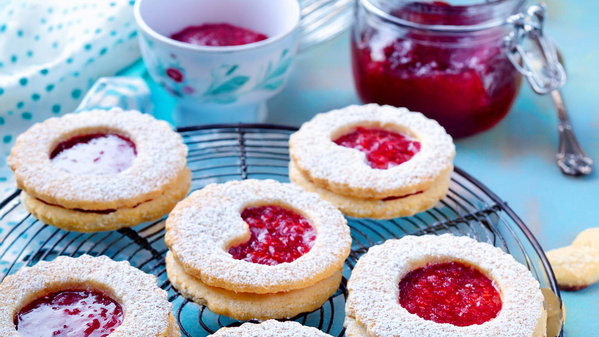 Das Bild zeigt Plätzchen mit Marmeladenfüllung und Puderzucker garniert, die auf einem Kuchengitter präsentiert werden. Im Hintergrund sind ein Glas mit Marmelade und eine Tasse zu sehen.