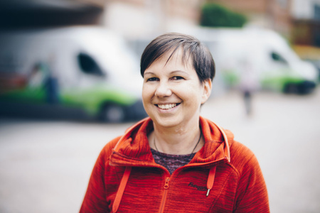 Eine Person in roter Jacke lächelt. Im Hintergrund stehen Lieferwagen.