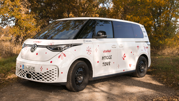 KI generiert: Das Bild zeigt einen weiß dekorierten Minivan mit der Aufschrift "HYGGE TOUR" auf einer ländlichen Straße, umgeben von herbstlichen Bäumen. Der Van trägt Motive, die eine gemütliche Atmosphäre vermitteln.