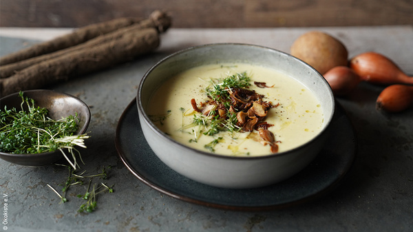 KI generiert: Eine Schüssel Suppe mit Kräutern, daneben Kresse, Wurzelgemüse und Zwiebeln. ©Ökokiste e. V.