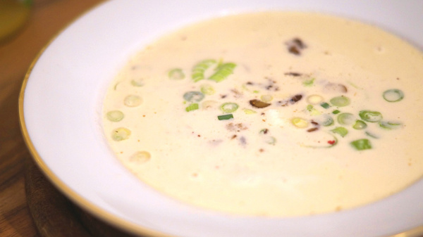 KI generiert: Das Bild zeigt eine Schüssel mit einer cremigen Suppe, die mit grünen Zwiebeln und Kräutern garniert ist. Die Suppe hat eine helle, beige Farbe und sieht reichhaltig aus.