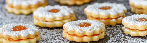 Das Bild zeigt mehrere übereinander gestapelte, blumenförmige Kekse, die mit Puderzucker bestäubt und dazwischen mit einer orangefarbenen Füllung versehen sind. Der Fokus liegt auf den sorgfältig arrangierten Keksen auf einer dunklen Oberfläche.