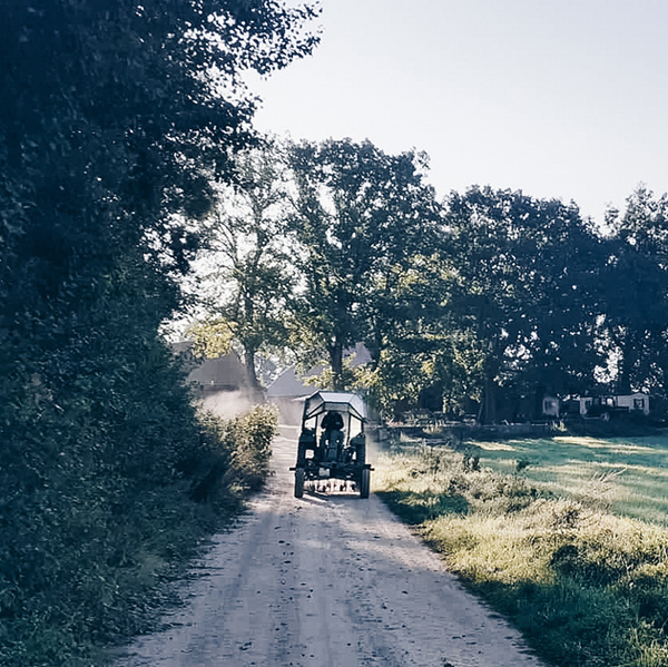Kleiner Trecker fährt Schotterweg entlang links Wald, rechts Wiese