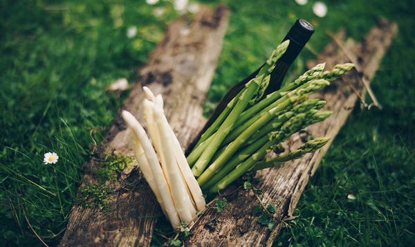 KI generiert: Weißer und grüner Spargel mit einer Weinflasche auf Holzbrett im Gras.