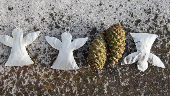 Drei Metallanhänger in Engelform und 2 geschlossene Kiefernzapfen liegen auf dem Boden mit leichtem Schneefalleinem 