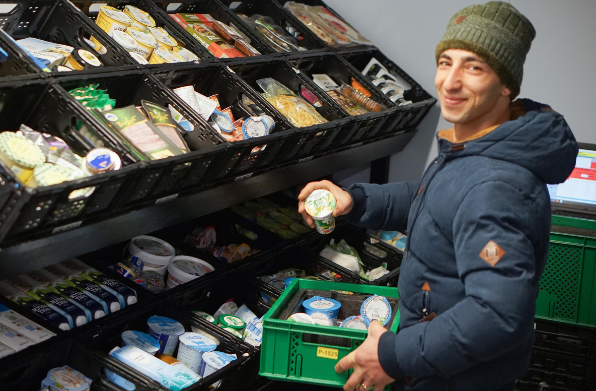 KI generiert: Ein Mann sortiert Lebensmittel in Regalfächer und hält eine grüne Kiste.