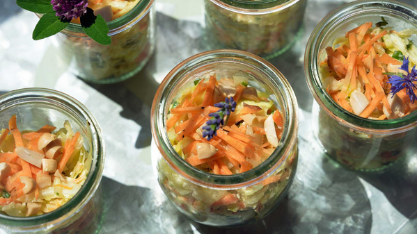 Kleine Gläser mit buntem Krautsalat, garniert mit essbaren Blüten und Kräutern, auf einem Tisch drapiert.