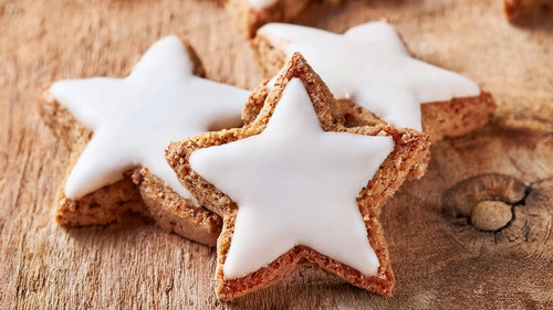 Das Bild zeigt sternförmige Plätzchen mit einer weißen Glasur auf einer Holzoberfläche. Die Plätzchen scheinen frisch gebacken und sind im Vordergrund fokussiert.