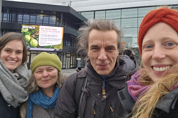 KI generiert: Vier lächelnde Personen posieren draußen vor einem Gebäude. Im Hintergrund steht auf einem Plakat "BIOFACH".