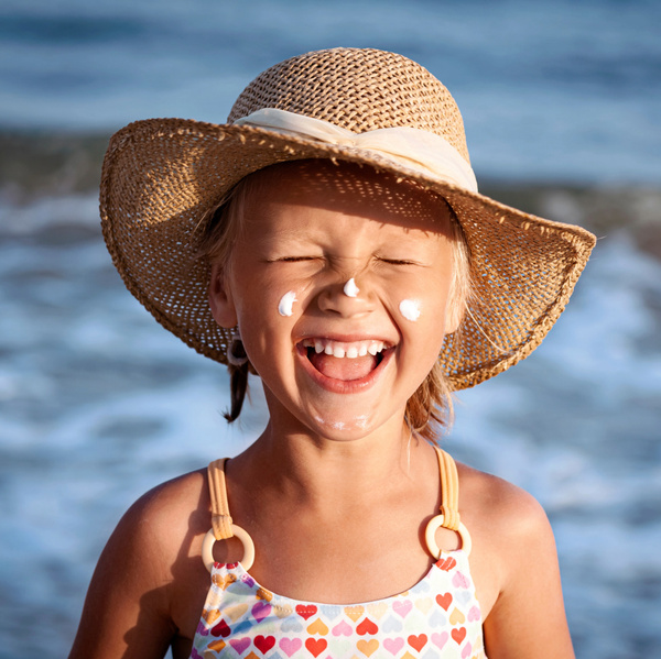 Mädchen mit Stroh-Sonnenhut und Sonnencremtupfen im Gesicht am Strand