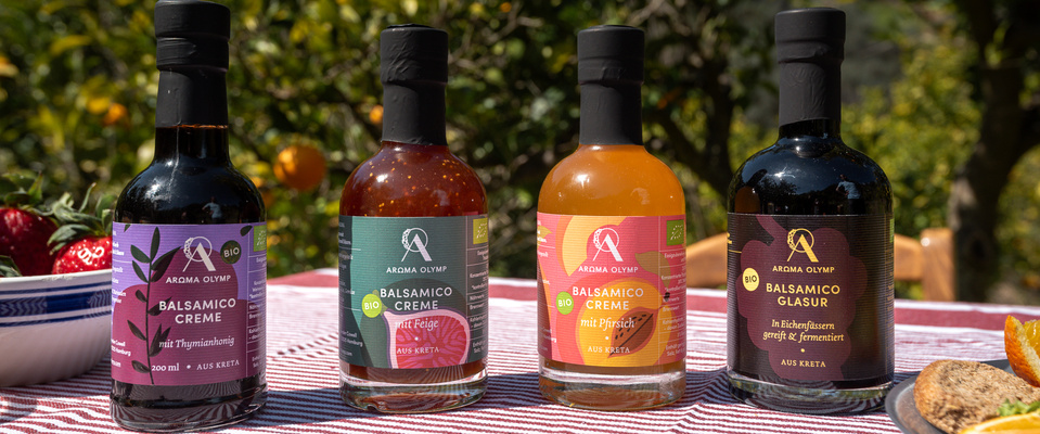 KI generiert: Vier Flaschen Balsamico mit verschiedenen Geschmacksrichtungen auf gestreifter Tischdecke. Text: „Aroma Olymp“.