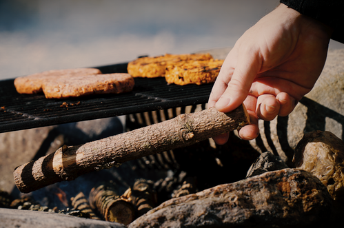 KI generiert: Hand legt einen Ast ins Feuer unter einem Grill mit Patties darauf.