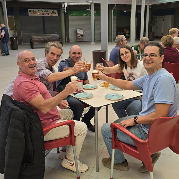 KI generiert: Eine Gruppe von fünf Personen sitzt an einem Tisch in einem hellen, überdachten Bereich und stößt mit Getränken an. Die Stimmung wirkt fröhlich und entspannt.