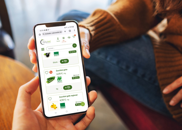 KI generiert: Jemand nutzt ein Smartphone für Online-Einkäufe bei "bioladen-salzwedel.de". Text: "Zucchini grün 4,49 €/kg".