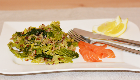 Das Bild zeigt einen Teller mit einer Portion Reis und Grünkohl, garniert mit rohem Lachs und Zitronenscheiben. Eine Gabel und ein Messer liegen daneben.