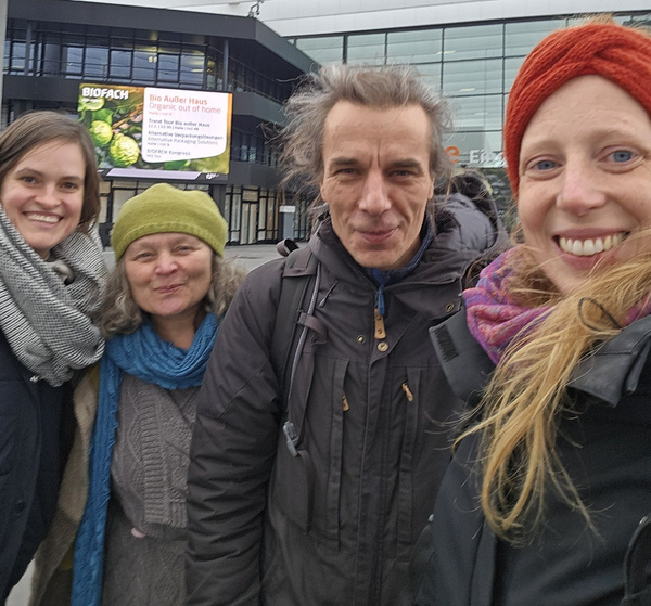 KI generiert: Vier Personen lächeln draußen in die Kamera. Text im Hintergrund: "BIOFACH Bio Außer Haus Organic out of home".