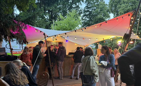 Im Bild ist ein überfüllter Open-Air-Stand unter einem bunt beleuchteten Zelt zu sehen.