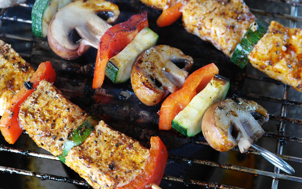 Gemüsespieße und mariniertes Fleisch werden auf einem Grill zubereitet. Keine Textbestandteile im Bild.