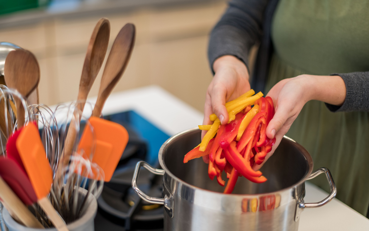 KI generiert: Das Bild zeigt eine Person, die geschnittene rote und gelbe Paprikastreifen in einem Topf in der Küche vorbereitet. Im Hintergrund sind verschiedene Küchenutensilien zu sehen.