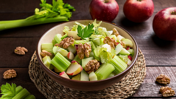 KI generiert: Ein Salat mit Sellerie, Äpfeln und Walnüssen in einer Schale auf einem Tisch, umgeben von Äpfeln und Sellerie.